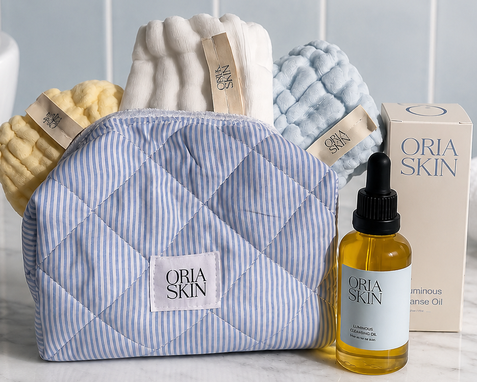 A quilted blue-and-white striped Oria Skin cosmetic pouch sits on a marble countertop with a bottle of facial oil and the product box beside it. Bath towels and a tiled wall are visible in the background.