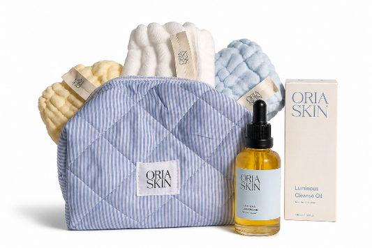 A skincare gift set from Oria Skin on a white background, featuring a blue and white striped quilted makeup bag, a bottle of Luminous Cleanse Oil, a cream box of the oil, and three textured washcloths in cream, white, and light blue.